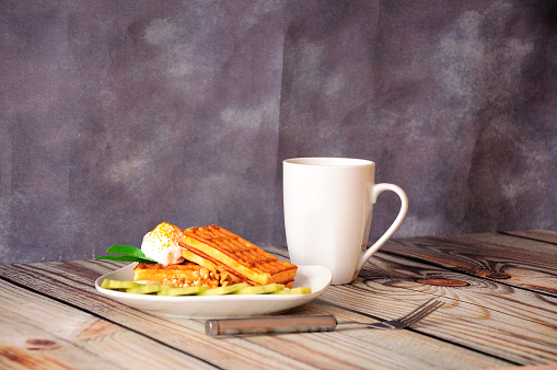 A plate of waffles with cream, slices of banana and kiwi, along with a cup of coffee, stand on a wooden table against a gray wall. Close-up.