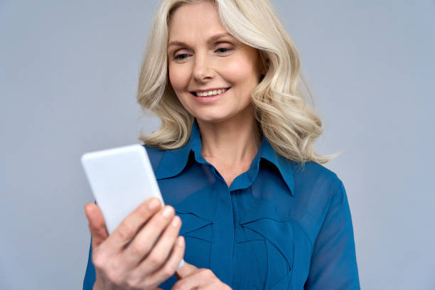 Happy smiling mature 50 years age blonde woman manager looking at cellphone using online mobile smartphone technology at device. Senior female leader professional shot isolated on gray background. feliz sonriente madura 50 años de edad rubia mujer gerente mirando el teléfono celular utilizando la tecnología de teléfonos inteligentes móviles en línea en el dispositivo. la líder profesional femenina de alto rango disparó aislada sobre fondo gr