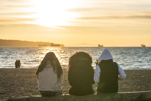 Three young girls enjoy the sunset at English Bay Beach, Vancouver City beautiful landscape. Concept of teenagers leisure.