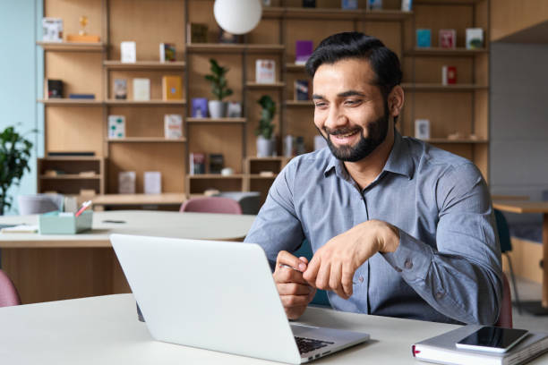 gelukkige glimlachende etnische indische zakenman die online virtuele vergaderingsoproep op laptop heeft die het schermzitting bij lijst in coworking creatief bureau bekijkt, professionele manager die met werknemers spreekt. - male employee office stockfoto's en -beelden