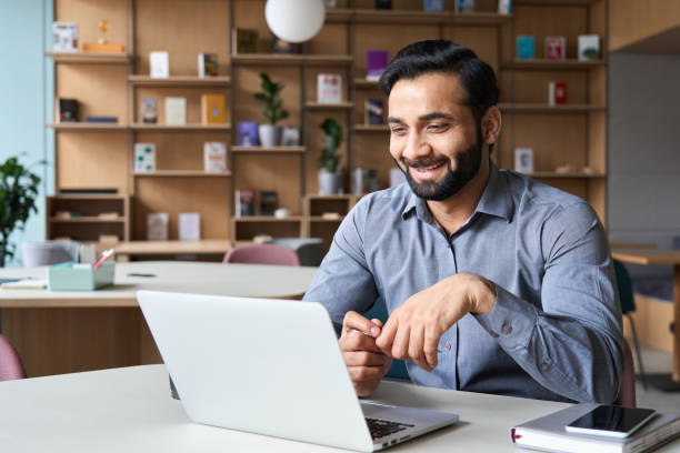 happy smiling ethnical indian businessman having online virtual meeting call on laptop looking at screen sitting at table in coworking creative office, professional manager talking to employees. - povo indiano imagens e fotografias de stock