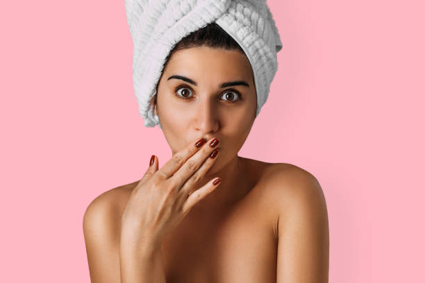 Portrait of a young attractive woman with a towel on her head on an isolated background. A girl with a towel on her head stands on a pink background, covering her mouth with her hand, close-up. stock photo