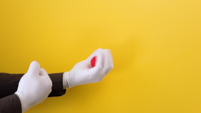 Male magician's hands in white gloves show the trick with a red foam ball. Yellow background. The concept of entertainment and illusions