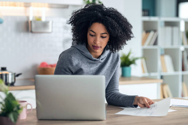 istock Afro business woman working with computer while consulting some invoices and documents in the kitchen at home. 1314926026