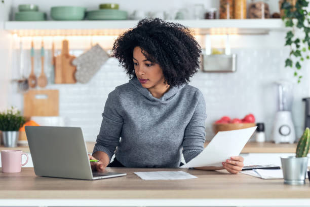 istock Afro business woman working with computer while consulting some invoices and documents in the kitchen at home. 1314926588