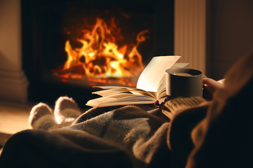 Woman with cup of drink and book near fireplace at home, closeup