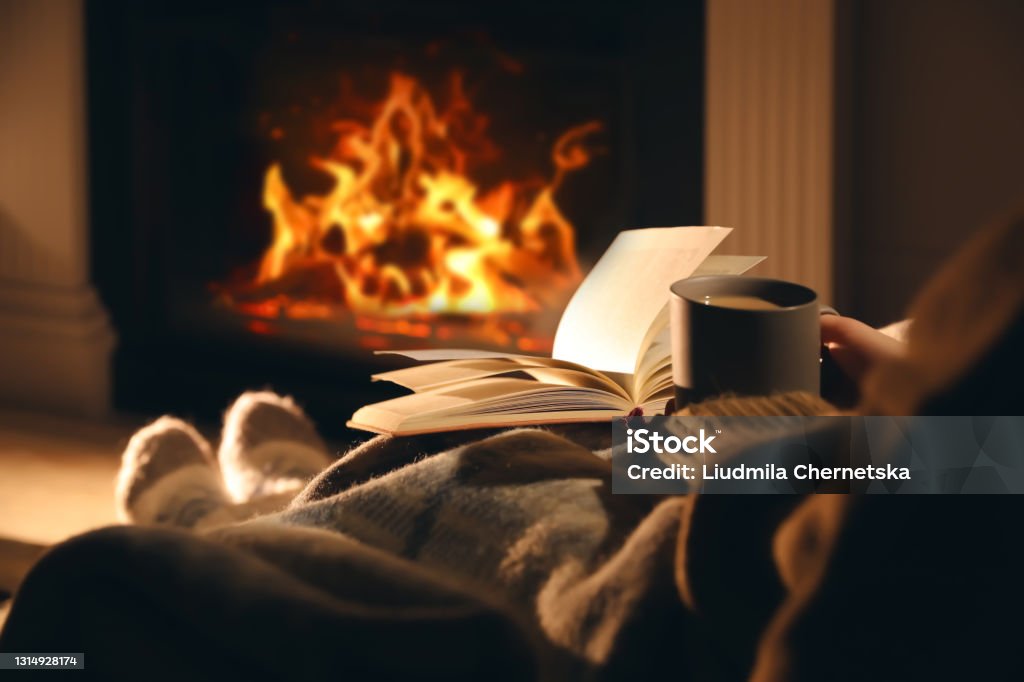 Woman with cup of drink and book near fireplace at home, closeup Fireplace Stock Photo