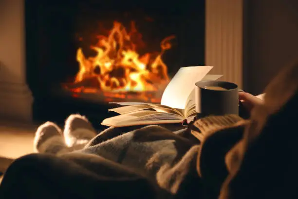 Photo of Woman with cup of drink and book near fireplace at home, closeup