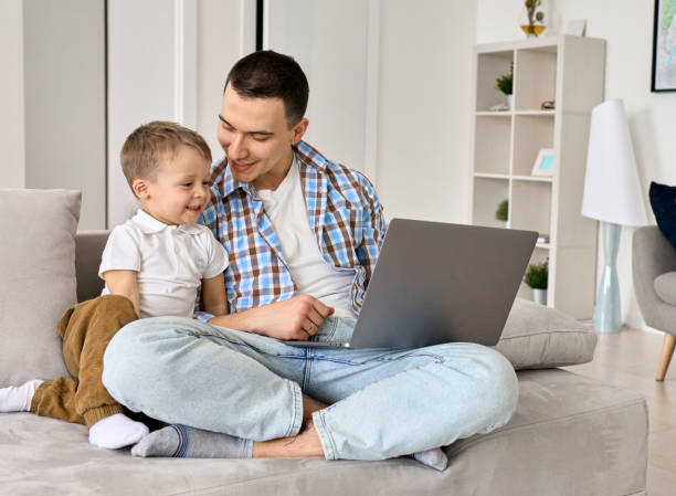 istock Happy young dad and cute small kid son using laptop computer at home. Smiling father distance learning or working online with toddler child, watching cartoons having fun spending time sitting on couch 1314936368