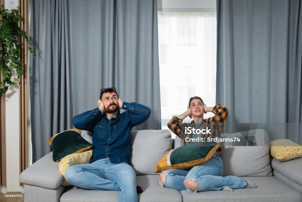 Young couple is sitting on a sofa in their apartment looking up and holding their hands to plug their ears as a neighbor upstairs is having a party and playing loud music or renovating the apartment Noise Stock Photo