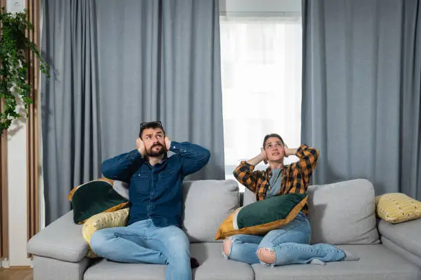 Photo of Young couple is sitting on a sofa in their apartment looking up and holding their hands to plug their ears as a neighbor upstairs is having a party and playing loud music or renovating the apartment