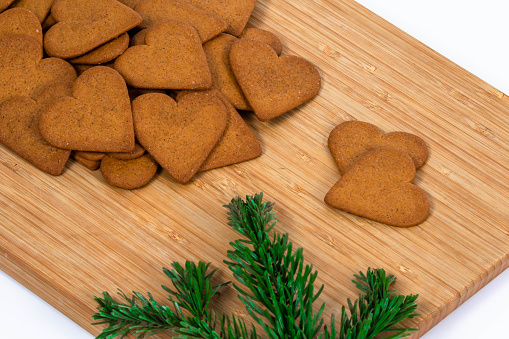 A pile of gingerbread hearts lying on a wooden cutting board, two hearts lying separately. A branch of green spruce tree lying in front of the cookies.