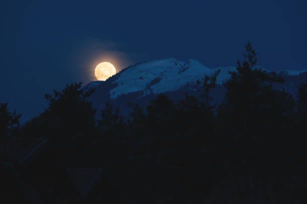 Supermoon setting dramatic behind snowy alpine mountain with evergreen forest, Austria