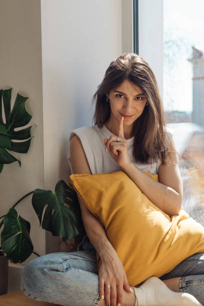 A dark-haired mixed-race girl sits on the windowsill and shows a gesture of silence.Home interior design.View of the city outside the window.The concept of relaxation and silence stock photo