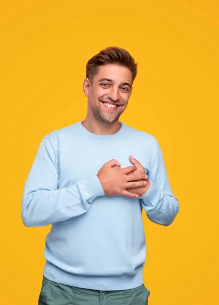 Flattered young man smiling for camera and holding hands on chest isolated on yellow background