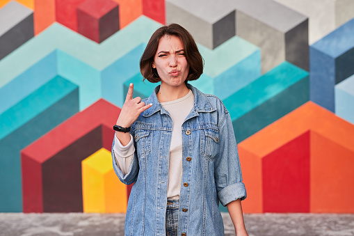 Modern millennial rebel female in denim jacket demonstrating horns sign while standing against wall with colorful geometric painting