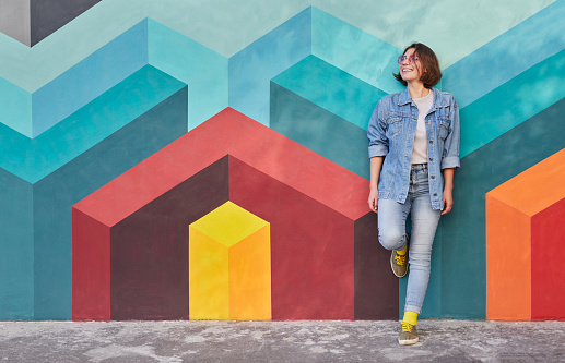 Cheerful young woman in denim clothes smiling and looking away while leaning on colorful wall with geometric graffiti in city