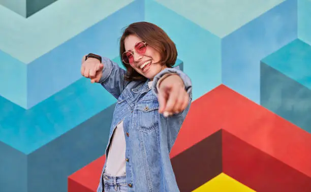 Happy hipster female in denim clothes and sunglasses pointing at camera in excitement on colorful background of graffiti