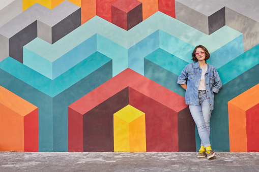 Full body female hipster in denim clothes leaning on colorful wall with geometric graffiti while resting on city street