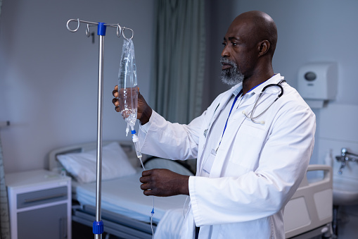 African american male doctor preparing iv drip bag on stand in hospital ward. medicine, health and healthcare services.