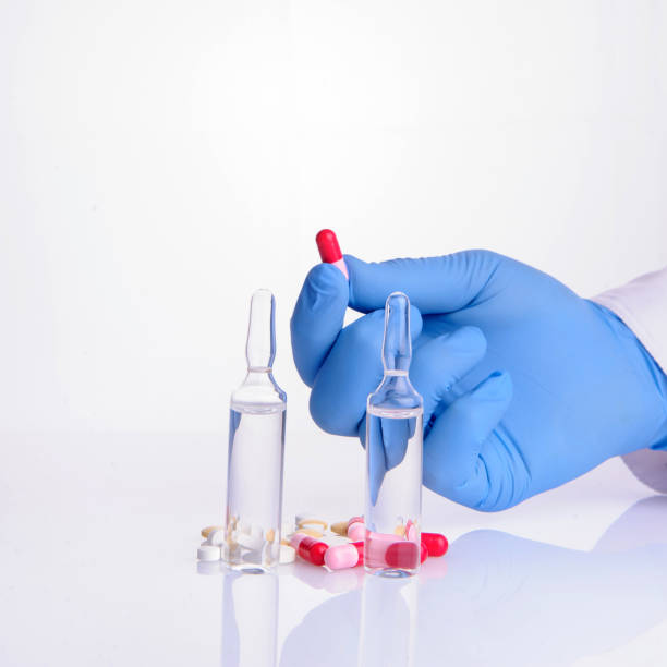 hand of a doctor holding a pill; two ampoules with medicine and other pills near it - surgical glove human hand holding capsule imagens e fotografias de stock