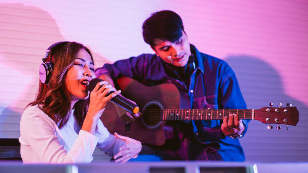 istock Pretty Asian female singer recording songs by using a studio microphone with male playing guitar in blue and red light. Performance and show in the music business. Duet session. 1314950823