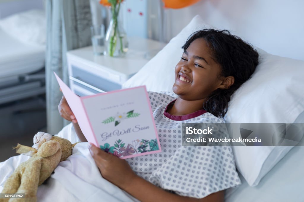 Smiling sick mixed race girl lying in hospital bed reading get well greetings card and holding teddy Smiling sick mixed race girl lying in hospital bed reading get well greetings card and holding teddy. medicine, health and healthcare services. Greeting Card Stock Photo