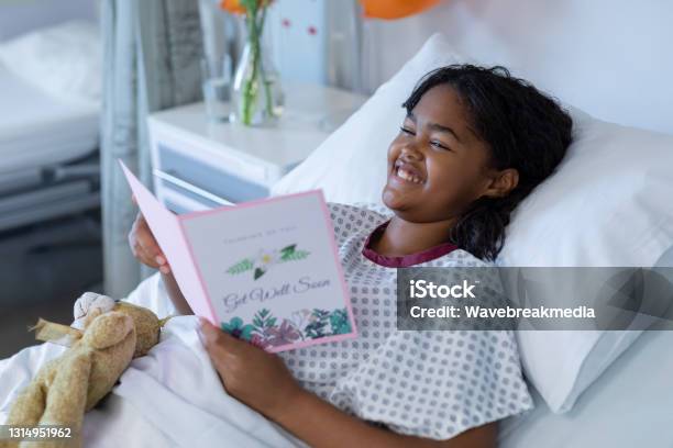 Smiling Sick Mixed Race Girl Lying In Hospital Bed Reading Get Well Greetings Card And Holding Teddy Stock Photo - Download Image Now