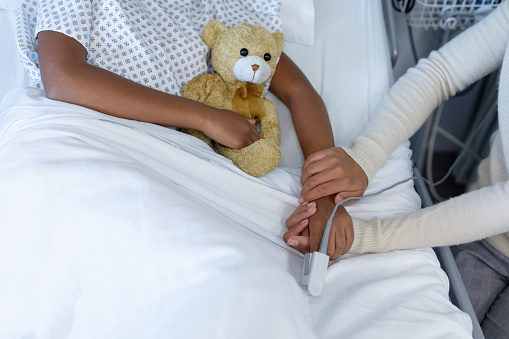 Midsection of mixed race mother and sick daughter in hospital, girl holding teddy bear. medicine, health and healthcare services during covid 19 coronavirus pandemic.