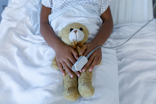 Midsection of sick mixed race girl in hospital bed wearing fingertip pulse oximeter holding teddy. medicine, health and healthcare services.