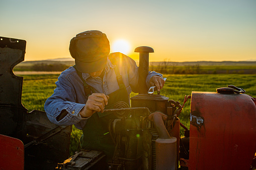 Older farmer repairing agricultural equipment on a farm during sunset