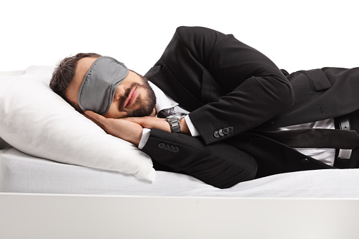 Businessman in a suit and tie sleeping on a bed and wearing a sleep mask isolated on white background
