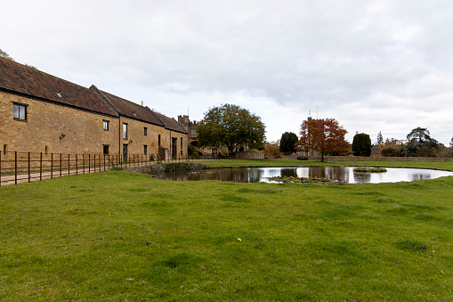Buildings and a Church behind the tree with reflection in a village pond in autumn. Single track road and field farmland grey sky