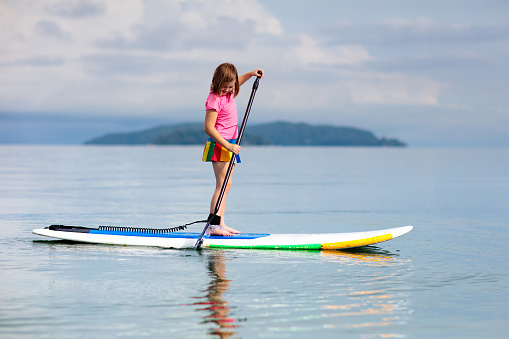 Child on stand up paddle board. Water fun and beach sport for kids. Healthy outdoor sports for summer vacation on tropical island. Holiday activity. Fit little girl training. Surfer exercising.