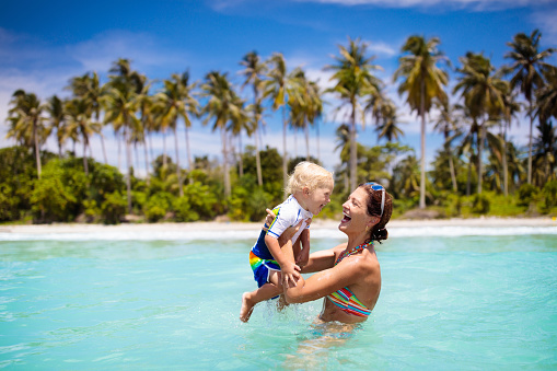 Mother and baby on tropical beach with coconut palm trees. Mom and little boy swimming and playing in water. Family summer vacation on exotic island. Travel with kids. Sun protection and swim wear.
