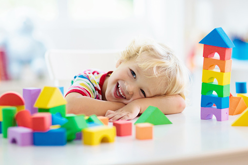 Kid playing with colorful toy blocks. Little boy building tower of block toys. Educational and creative toys and games for young children. Baby in white bedroom with rainbow bricks. Child at home.