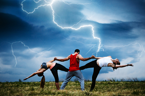 Three young Caucasian athlete people practicing acroyoga on meadow during storm.