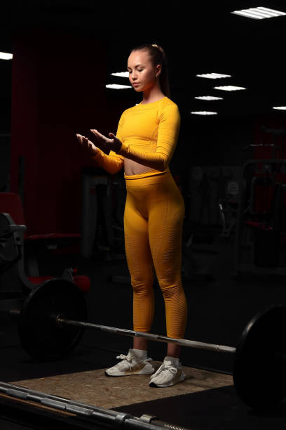 Young beautiful athletic girl in a yellow tight-fitting body tracksuit is about to do deadlift in the gym and looks at her palms with calluses. Fitness trainer shows exercises. European girl works out in the fitness center. Pulls the barbell to the chest stock photo