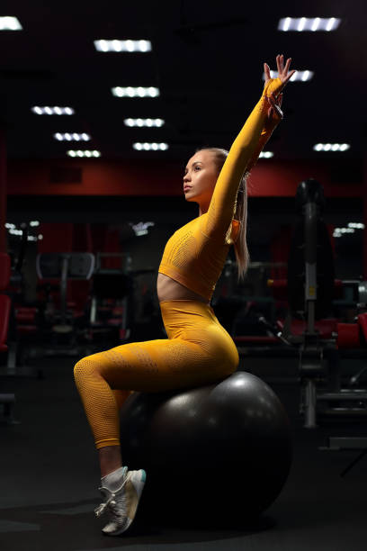 Young beautiful sportive girl in a yellow tight-fitting body tracksuit makes stretching with a fitness ball in the gym. Fitness trainer shows exercises. European girl workout stock photo