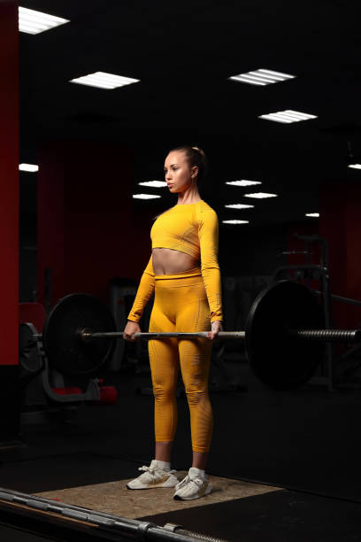 Young beautiful sportive girl in a yellow tight-fitting body tracksuit does deadlift in the gym. Fitness trainer shows exercises. European girl works out in the fitness center. Pulls the barbell to the chest stock photo