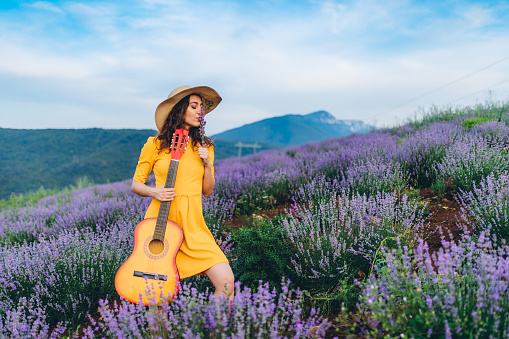 Young caucasian beautiful woman standing in lavender field with acoustic guitar.