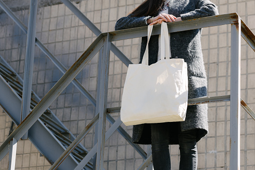 Woman holding white textile tote eco bag in urban area. Ecology or environment protection concept. White eco bag for mock up.