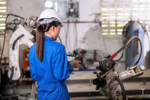 Engineer woman in uniform using remote control robotic arm machine to work in the factory. Worker setup program on the remote to do welding metal. Technology and industrial 4.0 concept.