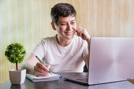 Brunette woman with short hair sits at a table in front of a laptop with a notebook and a pen. Woman smiling looking at the computer screen. Online courses, distance learning.