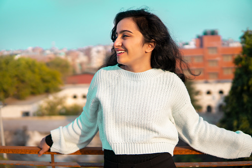 Beautiful happy Indian young woman standing near railing on rooftop. She is looking away and contemplating with a toothy smile and enjoying the fresh air at day time.