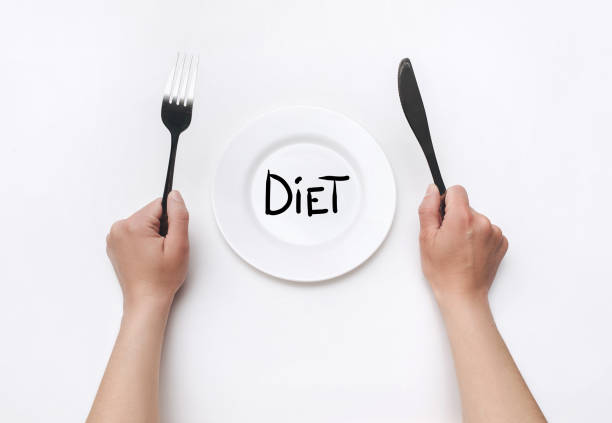 Female hands hold a fork and knife near a white plate on which the word Diet is written. The concept of a balanced diet, ration and medical fasting. Top view, white background, copy space. stock photo