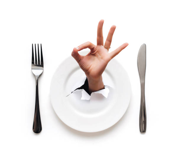 istock Plate,fork and knife lie on a white tablecloth.A hand appears through the plate in the torn hole of the paper with an OK gesture.The concept of a good restaurant and minimalist table setting, top view 1314994130