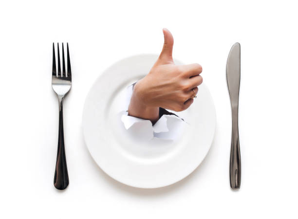 Plate,fork and knife lie on a white tablecloth.A hand appears through the plate in the torn hole of the paper with an Like gesture.The concept of a good restaurant and minimalist table setting, top view