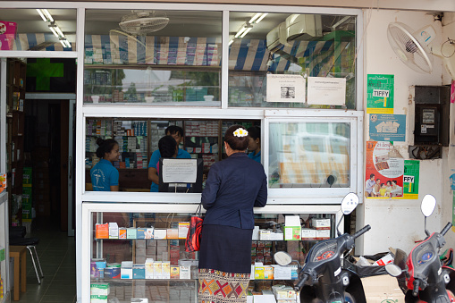 Laotian woman is standing at pharmacy in Vientiane. Woman is traditional dressed. She is standing at open window, At left side is open door. Inside are pharmacists.