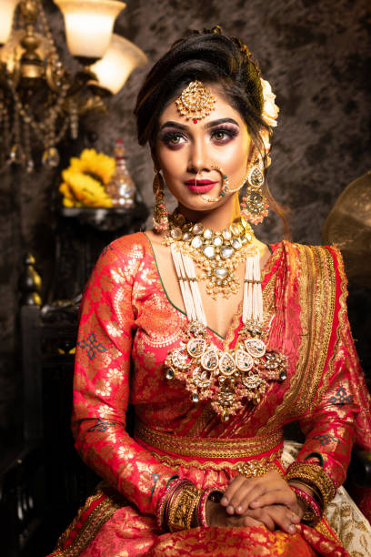 Stunning Indian bride in luxurious bridal costume with makeup and heavy jewellery is sitting in a chair in with classic vintage interior in studio lighting. Wedding lifestyle and fashion. stock photo